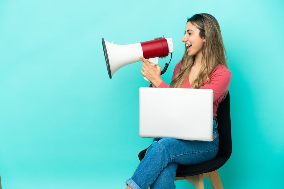 Young caucasian woman sitting chair with her pc isolated blue background shouting through megaphone